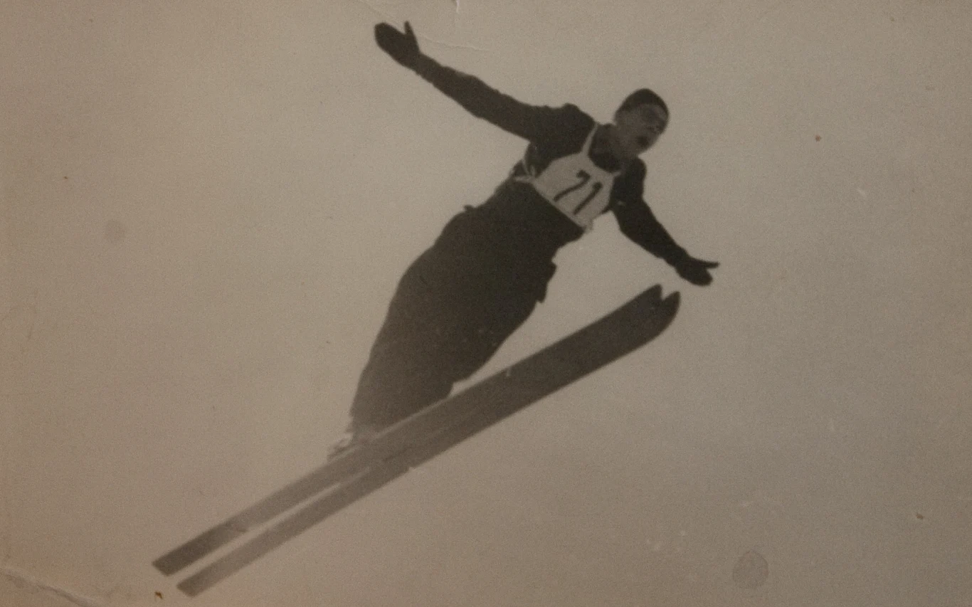 Skoczek narciarski w locie, ubrany w ciemny kombinezon i numer startowy 71, z ramionami szeroko rozłożonymi na boki. Tło jednolite, bez dodatkowych elementów.