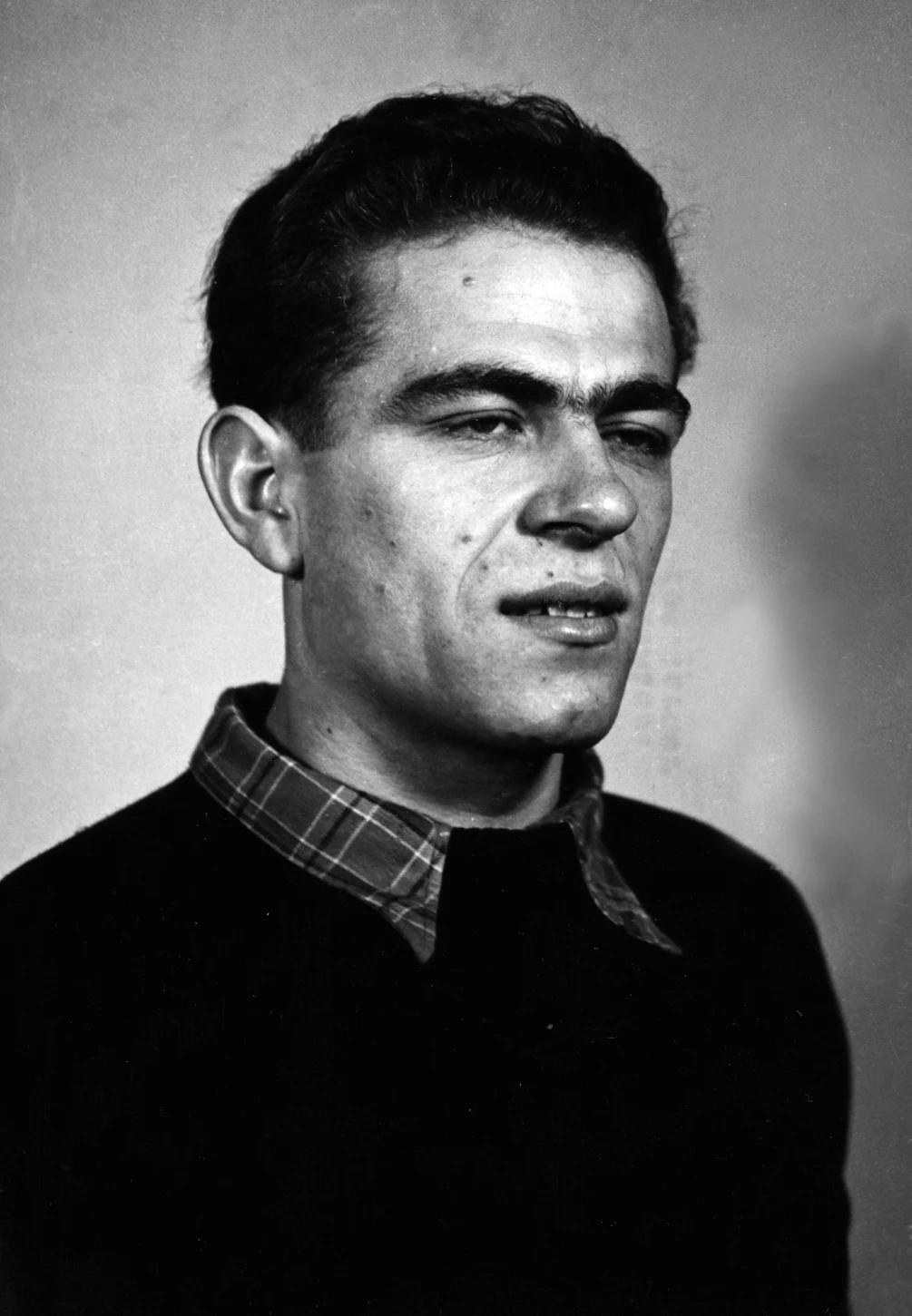 Mężczyzna o ciemnych włosach, z wyrazistymi rysami twarzy i lekkim zarostem, ubrany w ciemny sweter oraz koszulę w kratę, patrzący lekko w bok na neutralnym tle.