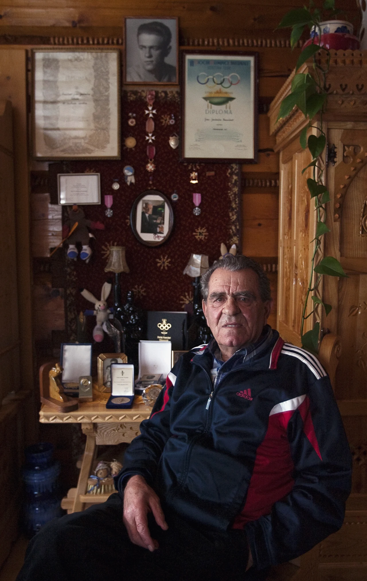 Starszy mężczyzna siedzący w drewnianym pomieszczeniu, ubrany w sportową kurtkę, otoczony medalami, odznaczeniami i pamiątkami wiszącymi na ścianie, powyżej widoczne fotografie i dyplomy nawiązujące do osiągnięć sportowych.