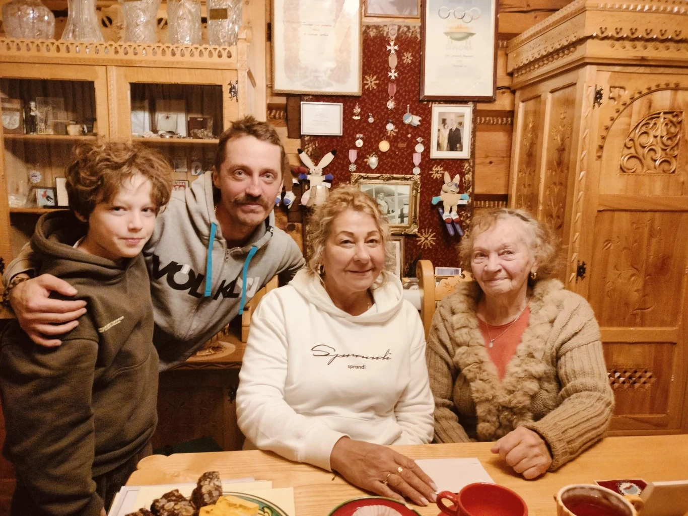 Cztery osoby w różnym wieku siedzą wspólnie przy stole w drewnianym, przytulnym wnętrzu. Na stole leżą filiżanki, talerze z jedzeniem i dokumenty. W tle widoczne są drewniane meble, półki z naczyniami i ściana ozdobiona dekoracjami oraz dyplomami.