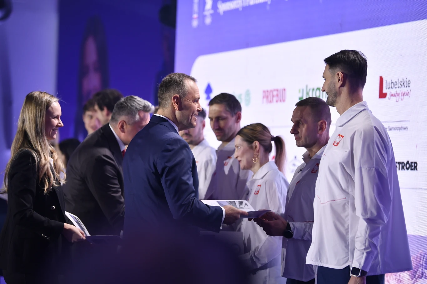 Grupa osób ubranych w oficjalne stroje sportowe otrzymuje nagrody lub dyplomy od przedstawicieli podczas oficjalnej uroczystości na tle tablicy z logotypami partnerów i sponsorów.