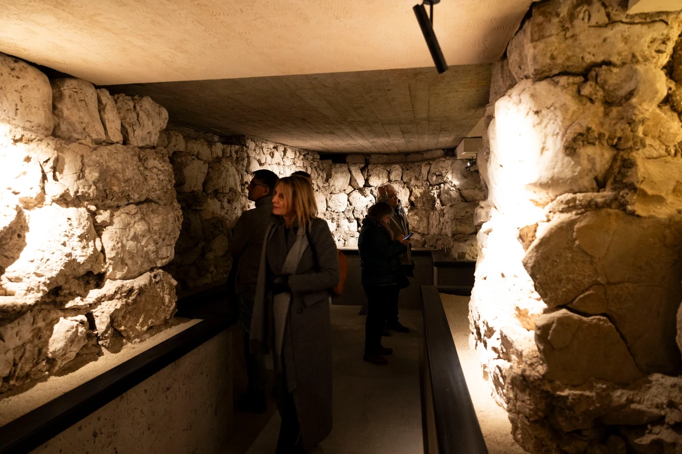 Grupa osób zwiedzająca podziemne korytarze otoczone masywnymi kamiennymi ścianami, przy sztucznym oświetleniu podkreślającym strukturę murów.
