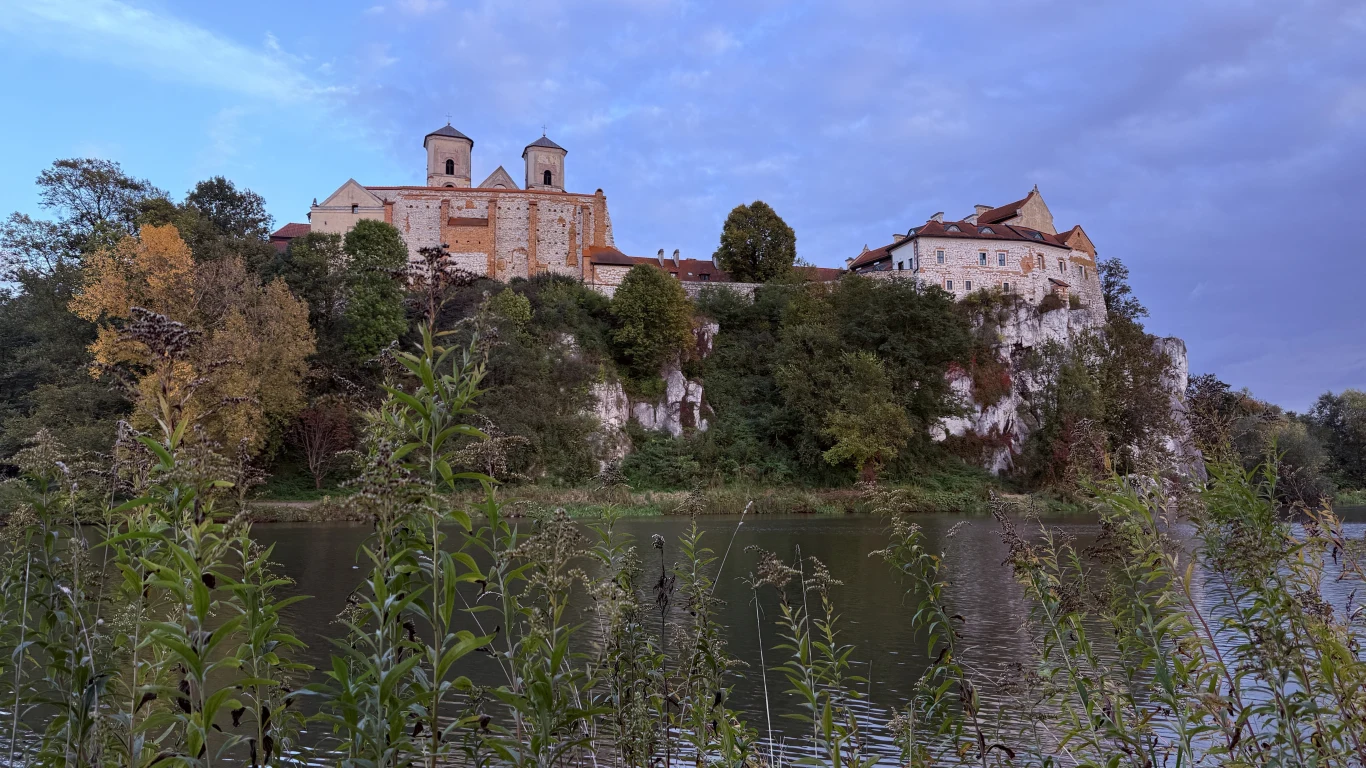 Romantyczny klasztor wzniesiony na wapiennym wzgórzu otoczony bujną zielenią, u podnóża którego płynie spokojna rzeka. W tle niebo z delikatnymi chmurami, otoczenie zdominowane przez naturalną roślinność.