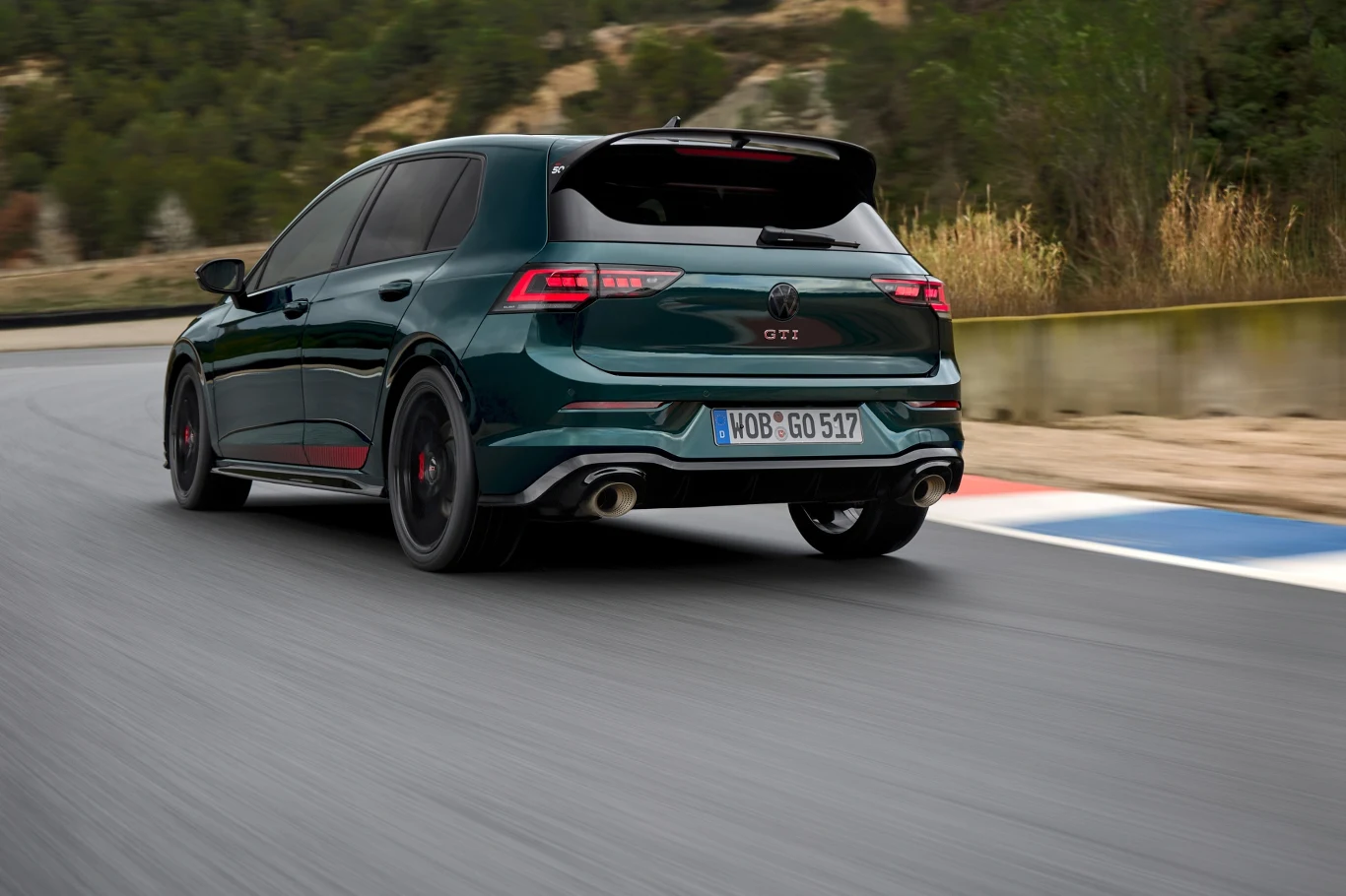 Ciemnozielony samochód sportowy Volkswagen Golf GTI w ruchu na torze wyścigowym, widok od tyłu, otoczenie stanowi asfaltowa nawierzchnia oraz zieleń w tle.