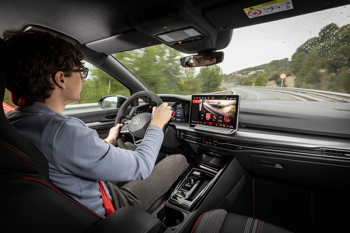 Mężczyzna prowadzi nowoczesny samochód po pustej drodze, skupiając uwagę na trasie. Wnętrze auta wyposażone jest w cyfrowy wyświetlacz, nowoczesny panel sterowania oraz wygodne fotele.