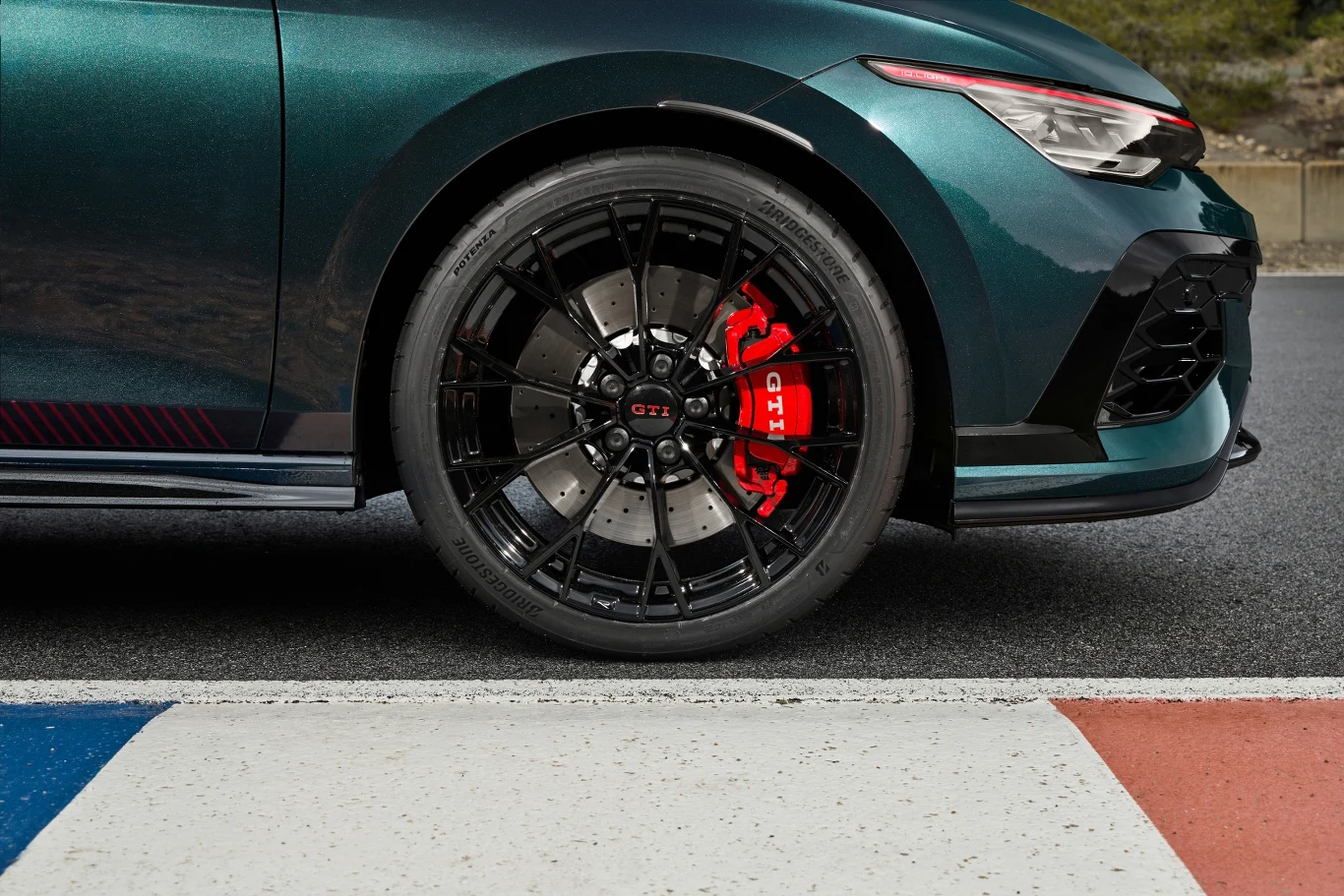 Zbliżenie na przednią część nowoczesnego sportowego samochodu, czarny aluminiowy felg z czerwonym zaciskiem hamulcowym GTI, wyeksponowany na tle toru wyścigowego z widocznymi kolorowymi liniami.