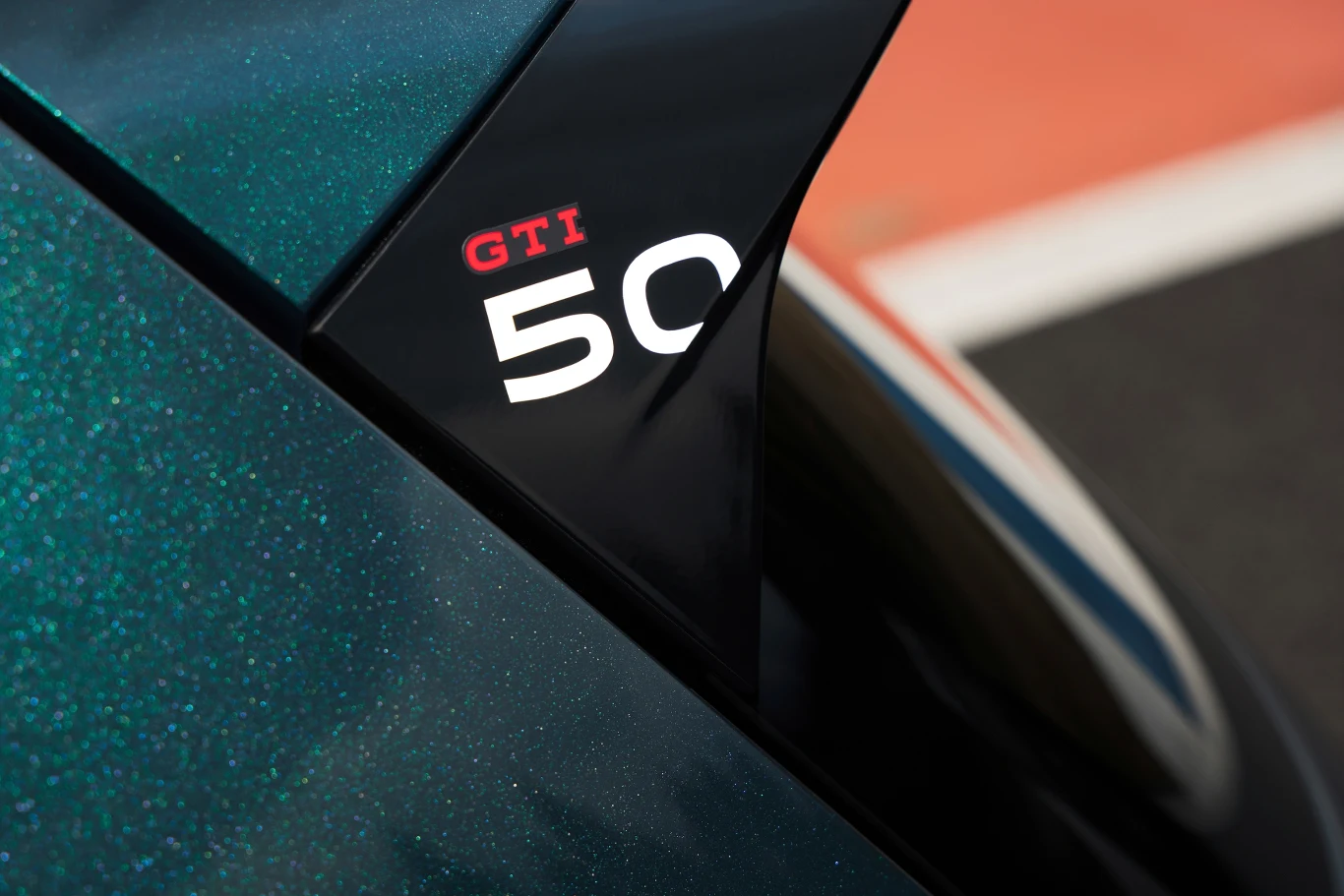 Czarny detal samochodu z widocznym napisem GTI 50 na błyszczącej powierzchni nadwozia, obok fragment drogi z białą linią.