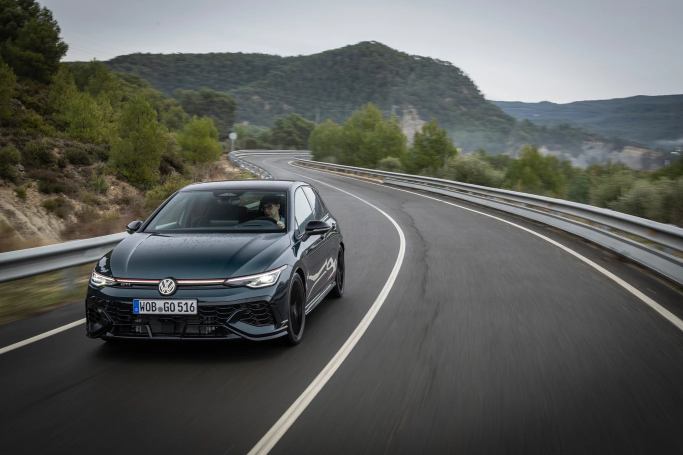 Czarny samochód Volkswagen Golf jedzie pustą, asfaltową drogą wijącą się przez zielone, pagórkowate tereny, otoczony barierkami i drzewami, z górami widocznymi na dalszym planie pod pochmurnym niebem.