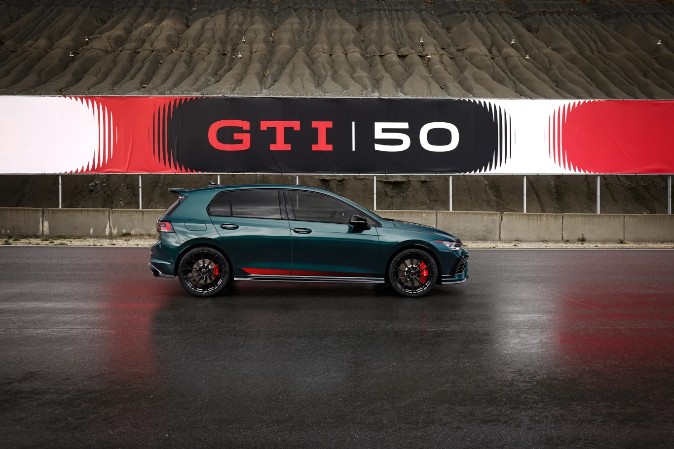 Sportowy samochód Volkswagen Golf GTI zaparkowany na mokrej nawierzchni na tle dużego bannera z napisem GTI 50 oraz charakterystyczną grafiką w kolorach czarnym, czerwonym i białym, z widocznym wzgórzem w tle.