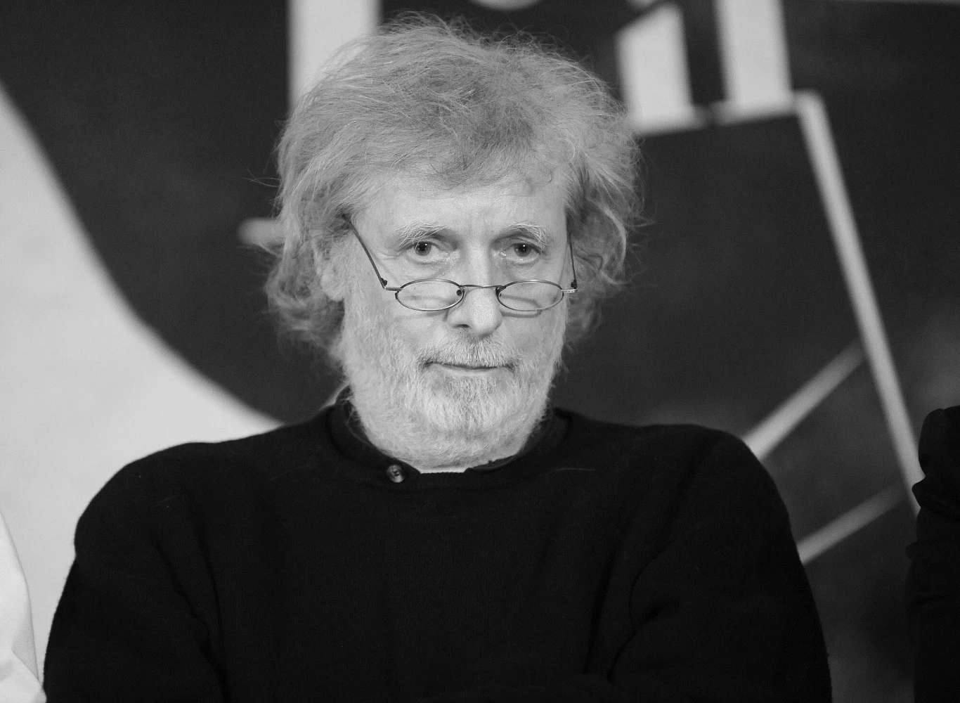 Wojciech Michniewski miał 78 lat Starszy mężczyzna z siwymi włosami i brodą, w okularach, ubrany w ciemny sweter. Postać znajduje się na tle graficznym lub plakatu. Twarz wyraża powagę i skupienie.