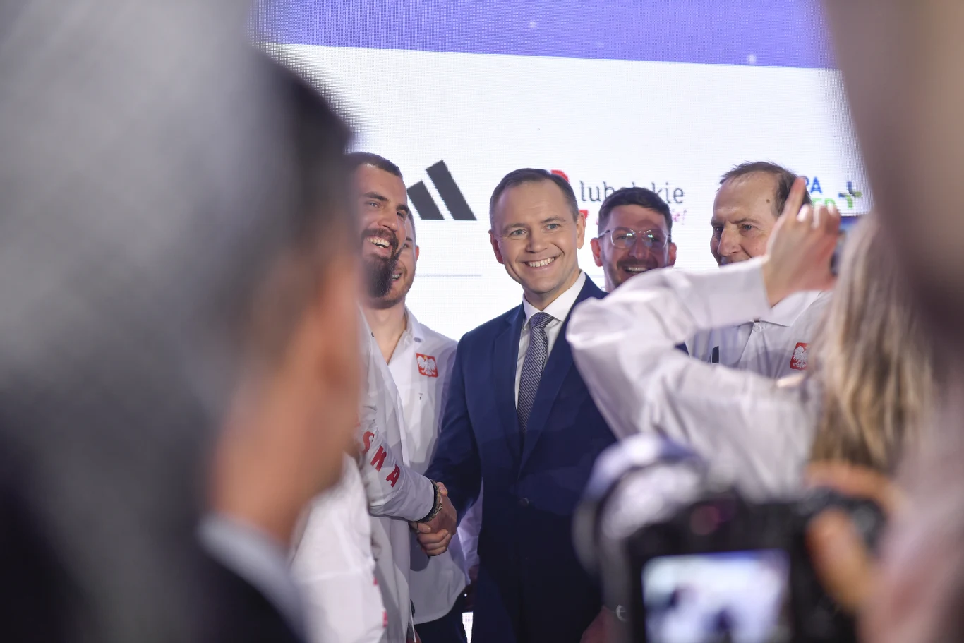 Grupa mężczyzn w biznesowych garniturach i koszulach sportowych z logo Polski uśmiecha się, ściskając sobie dłonie podczas oficjalnego wydarzenia, w tle widoczne są logotypy sponsorów.