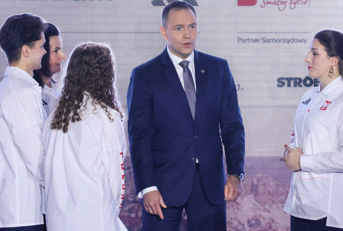 Mężczyzna w eleganckim garniturze stoi w centrum, otoczony przez grupę czterech młodych osób w białych sportowych kurtkach z polskimi emblematami, wszyscy znajdują się na tle z logotypami partnerów i hasłami.