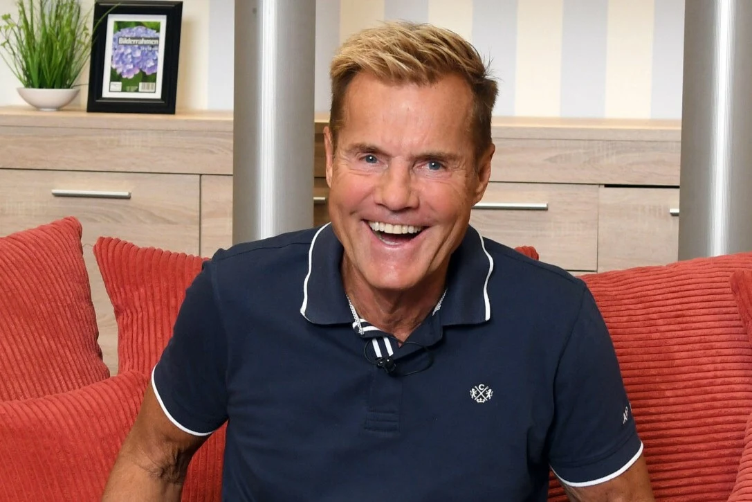 Tak wygląda obecnie 71-letni Dieter Bohlen Uśmiechnięty mężczyzna w granatowej koszulce polo siedzący na czerwonej sofie. Za nim znajduje się drewniana komoda, poduszki oraz roślina doniczkowa i zdjęcie w ramce.