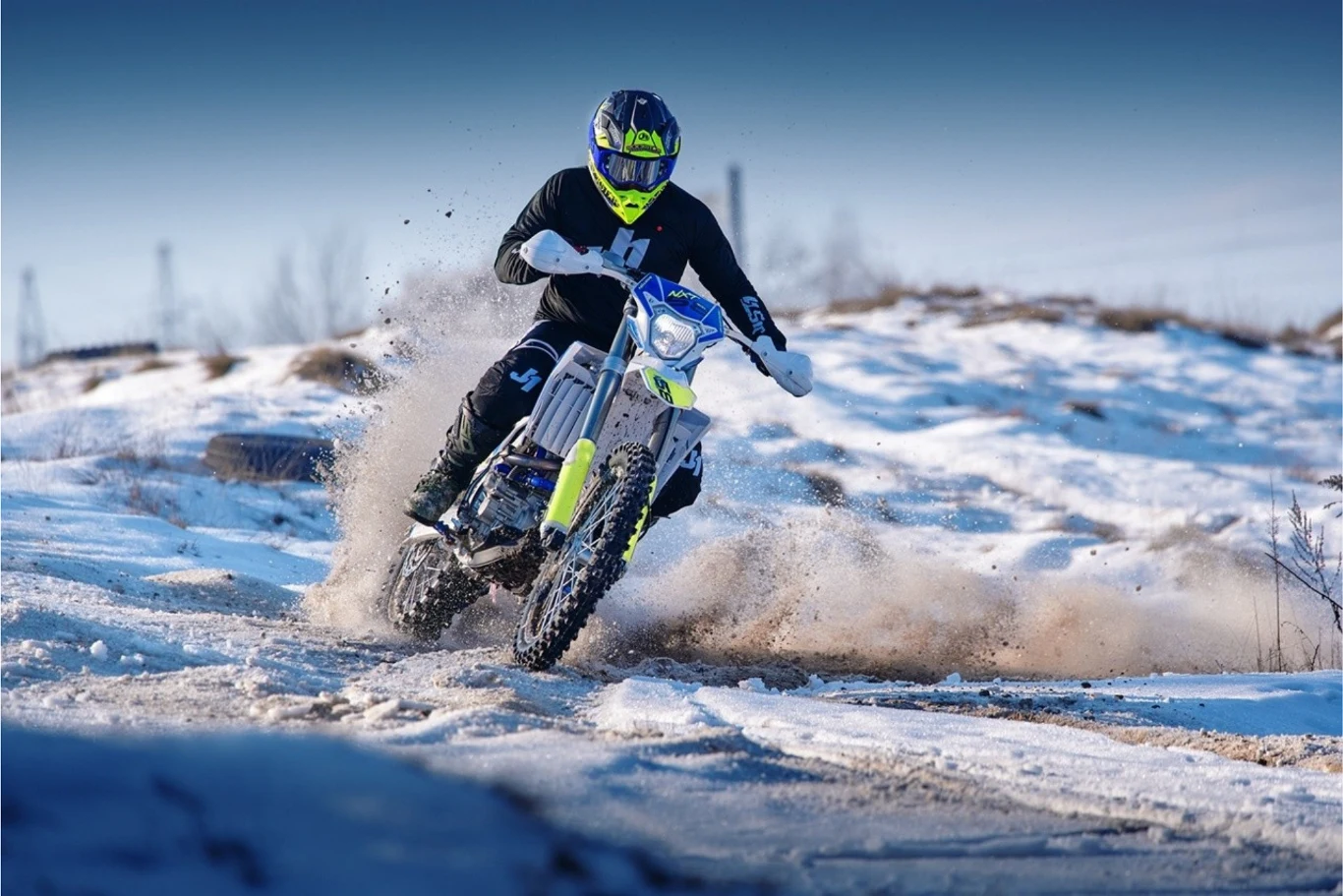 Barton prezentuje odświeżoną serię NXT na rok 2026. Motocyklista w niebiesko-czarnym stroju i żółtym kasku jedzie terenowym motocyklem przez ośnieżoną, błotnistą trasę, wzbijając za sobą rozpryskujący się śnieg