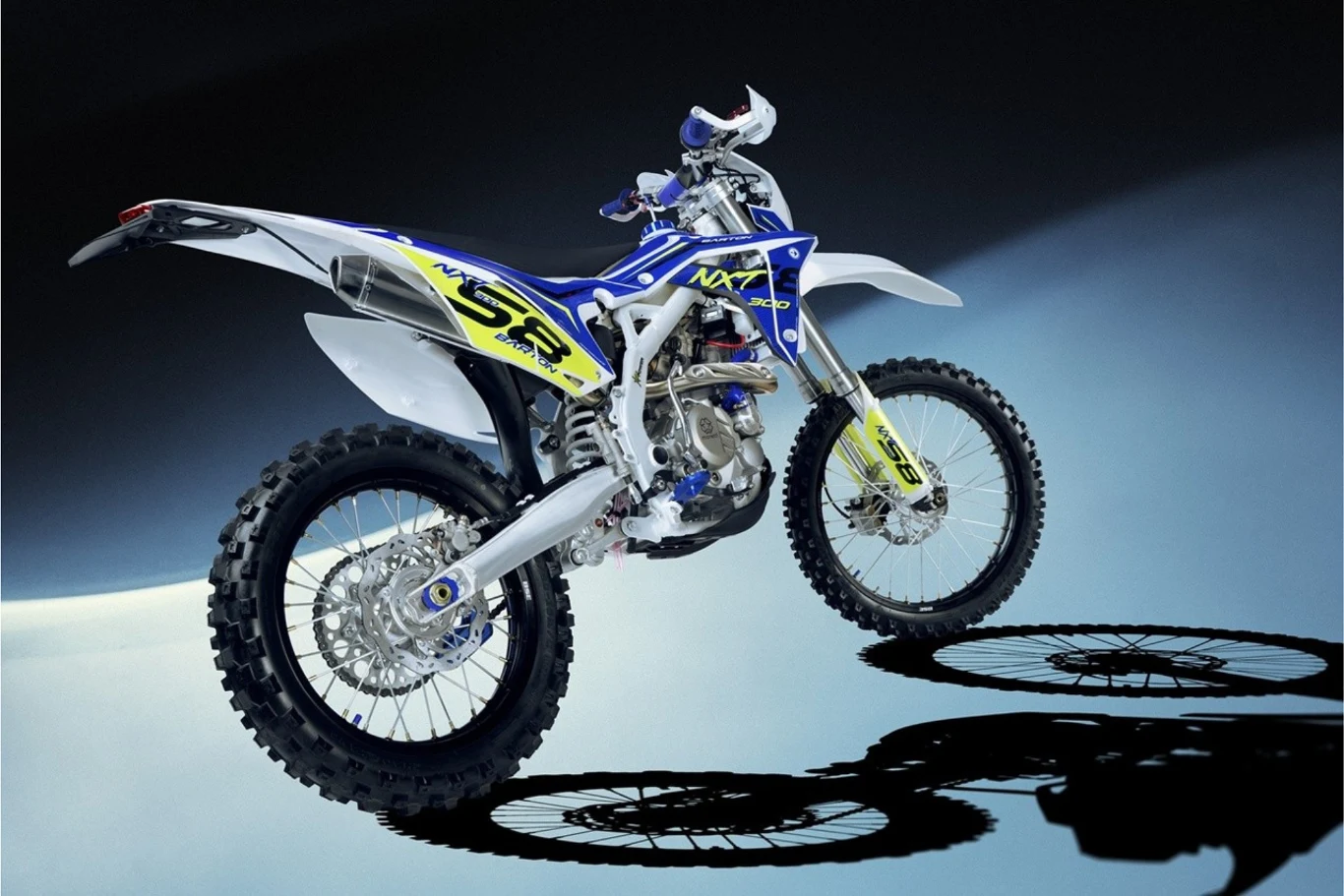 Motocykl terenowy typu enduro w sportowej kolorystyce z niebieskimi i żółtymi akcentami, ustawiony na jasnym tle, rzucający wyraźny cień na podłoże. Widoczne detale takie jak koła z grubym bieżnikiem, rama oraz sportowe oznaczenia na karoserii.