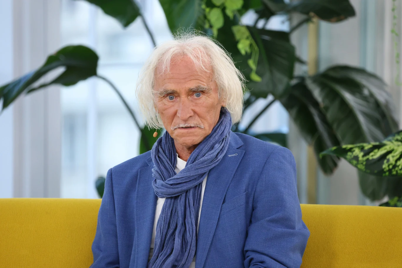 Jerzy Kryszak Starszy mężczyzna z białymi włosami ubrany w niebieską marynarkę i szalik siedzi na żółtej sofie, w tle duże zielone rośliny doniczkowe oraz jasne wnętrze.