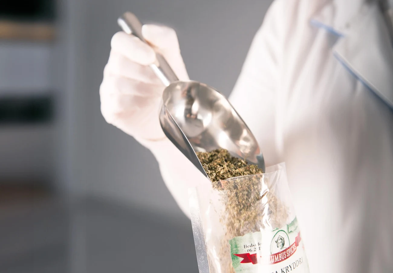 Ręka w jednorazowej rękawiczce wsypuje suszone zioła do przezroczystej torebki za pomocą metalowej łopatki, na opakowaniu widoczne etykiety z informacjami o produkcie.