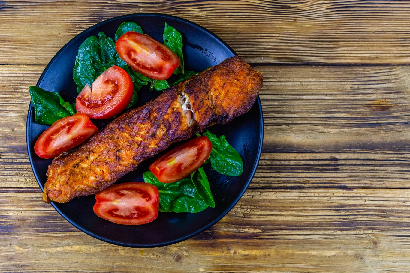 Grillowany filet z ryby podany na czarnym talerzu ze świeżymi liśćmi szpinaku i plasterkami pomidora, wszystko na tle drewnianego stołu.