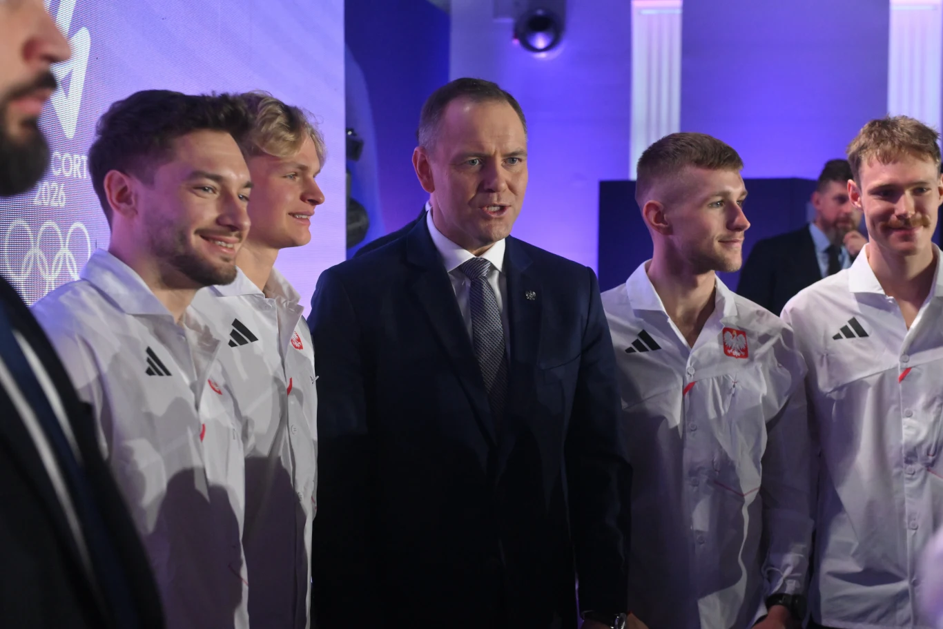 Grupa młodych mężczyzn ubranych w jednakowe, białe sportowe kurtki z godłem Polski stoi obok mężczyzny w garniturze, wszyscy uśmiechnięci, sfotografowani w dobrze oświetlonym, oficjalnym wnętrzu z widocznym elementem związanym z igrzyskami olimpijskimi.