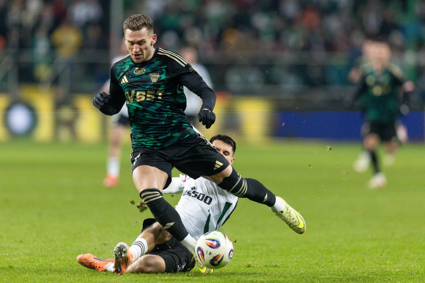 Dwóch piłkarzy walczy o piłkę podczas meczu, jeden z nich wykonuje wślizg, próbując zatrzymać rywala w dynamicznej akcji na boisku.