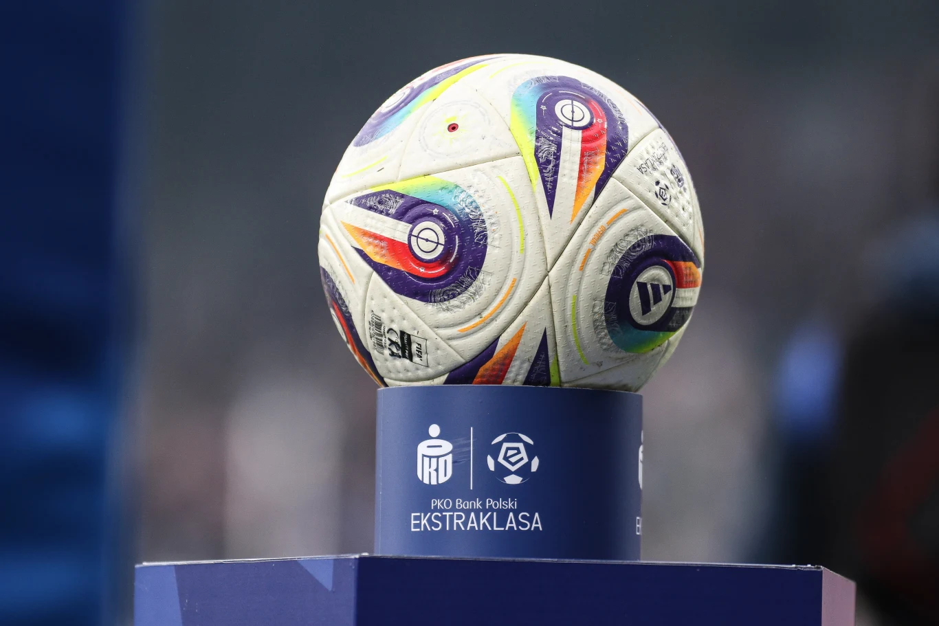 Oficjalna piłka meczowa z kolorowymi wzorami umieszczona na niebieskim postumencie z logotypami PKO Bank Polski Ekstraklasa na tle rozmytego stadionu.