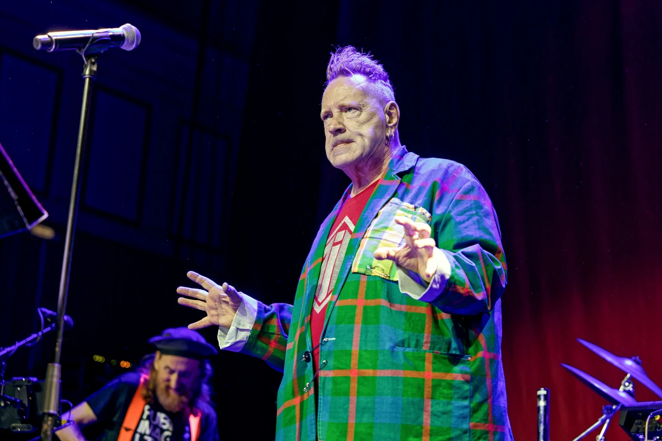 Mężczyzna w kolorowej marynarce w kratę i czerwonej koszulce stoi na scenie przy mikrofonie, w tle widoczny kolejny muzyk i część perkusji, scena podświetlona niebieskim i fioletowym światłem.