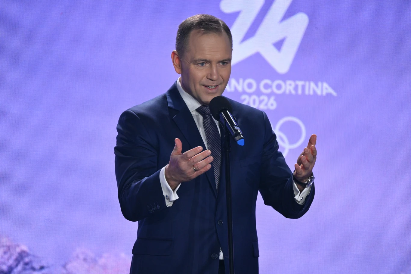 Mężczyzna w garniturze przemawia do mikrofonu na tle fioletowej ścianki z logo Zimowych Igrzysk Olimpijskich Milano Cortina 2026.