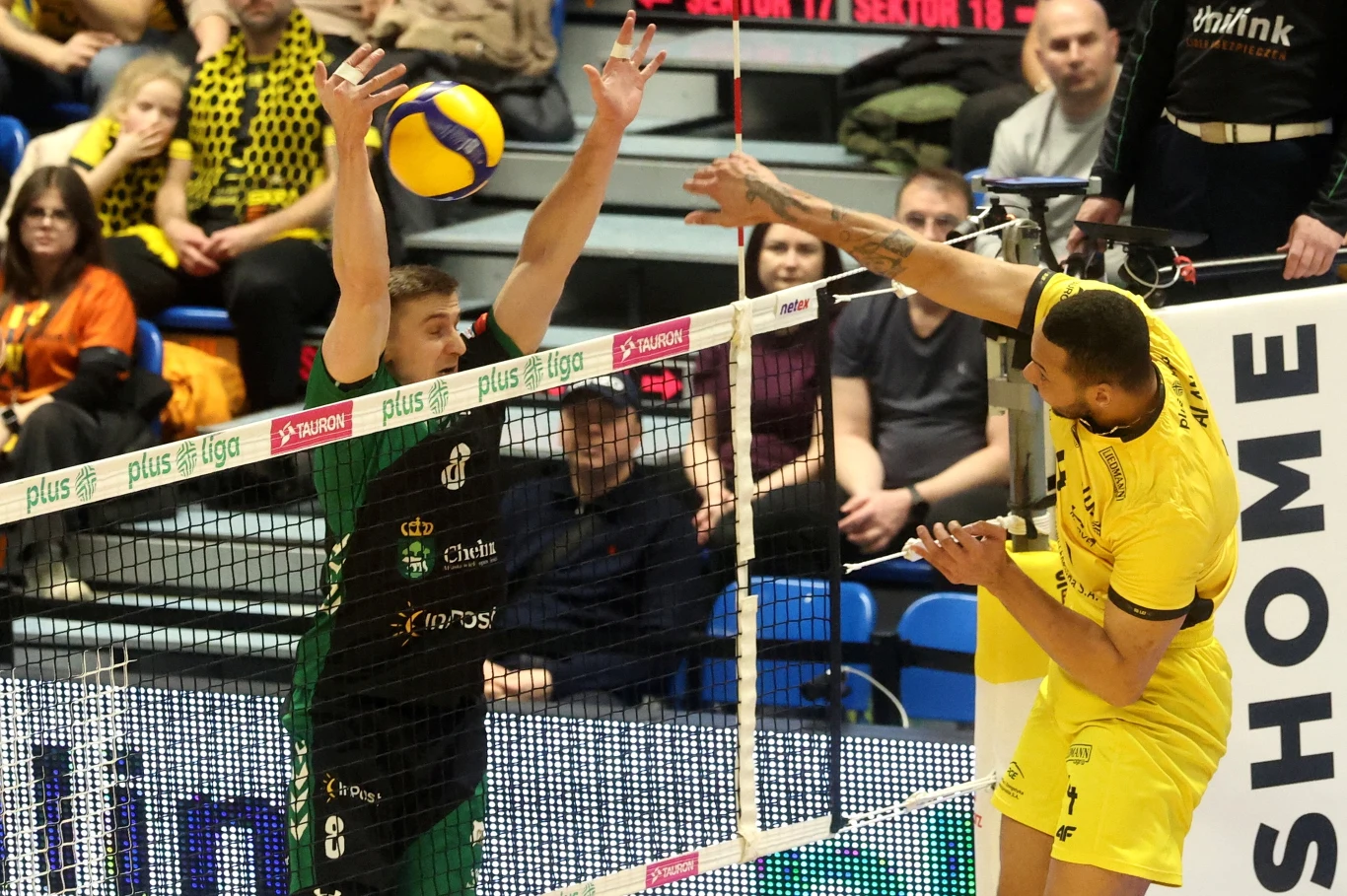 Kadr z meczu PGE GiEK Skra Bełchatów - InPost ChKS Chełm: Paweł Rusin kontra Alan Souza Dwóch siatkarzy podczas dynamicznej akcji przy siatce, jeden z nich blokuje, a drugi ścina piłkę tenisową, w tle widownia uważnie obserwuje mecz.