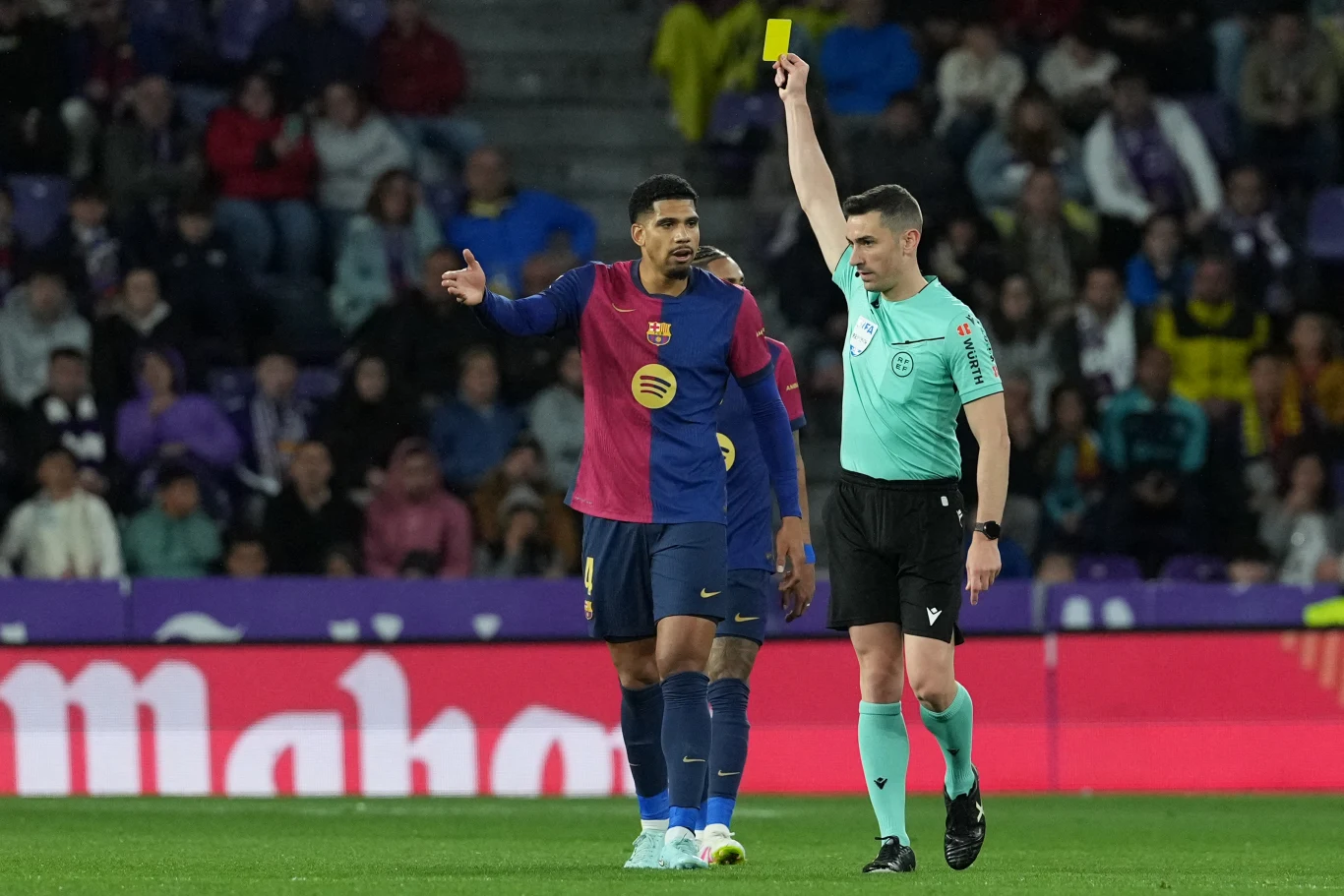 Sędzia piłkarski unosi rękę, pokazując żółtą kartkę zawodnikowi drużyny FC Barcelona w trakcie meczu, a zawodnik gestykuluje niezadowolony z decyzji. W tle widoczni kibice na trybunach stadionu.