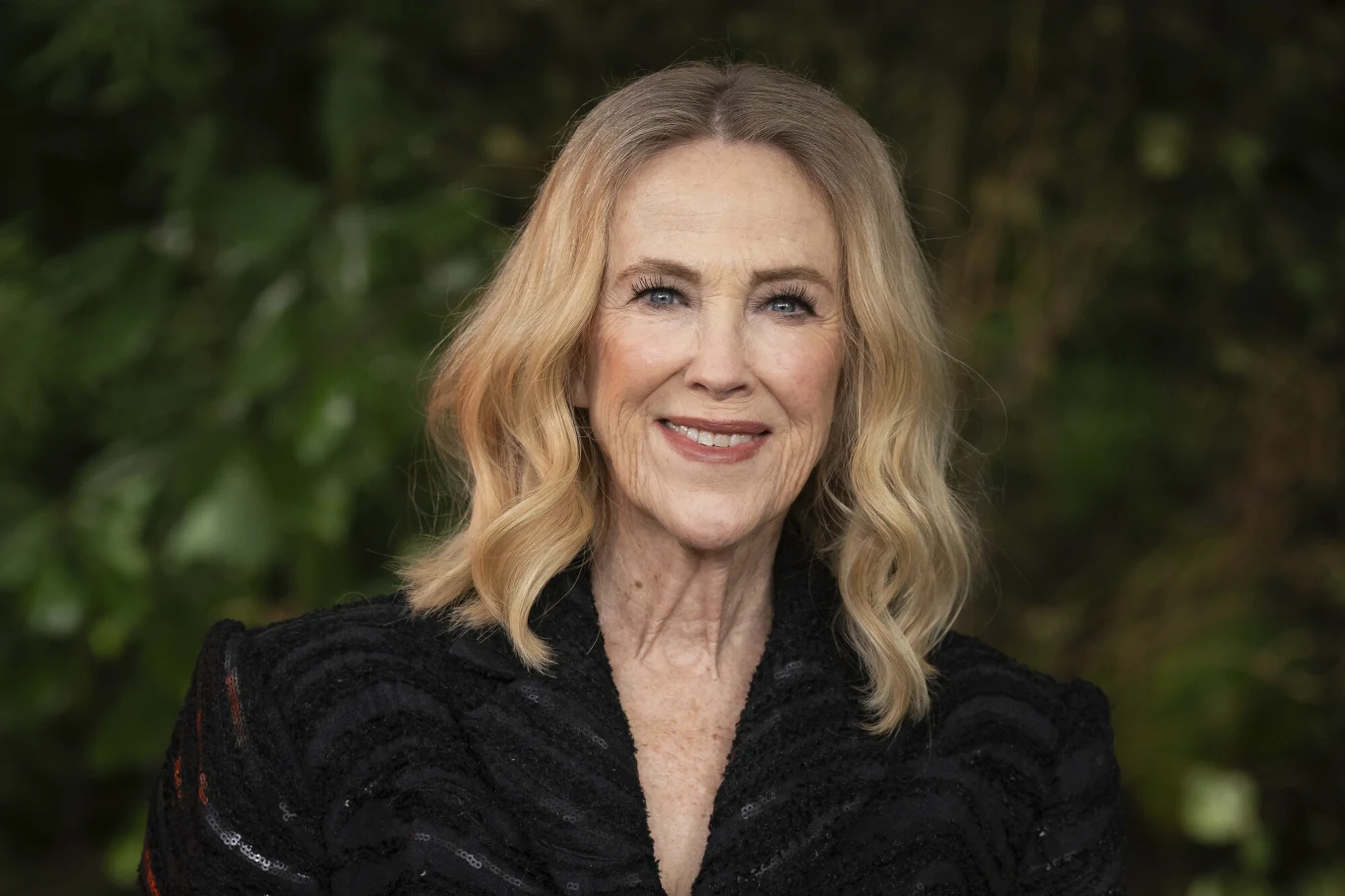 Catherine O'Hara Catherine O'Hara w eleganckim czarnym stroju, uśmiechnięta na tle zieleni.