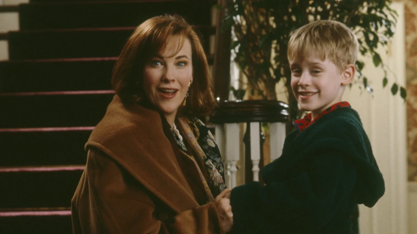 "Kevin sam w domu" Uśmiechnięta kobieta ubrana w brązowy płaszcz trzyma chłopca w zielonej kurtce za ręce, stojąc razem przy schodach w domowym wnętrzu