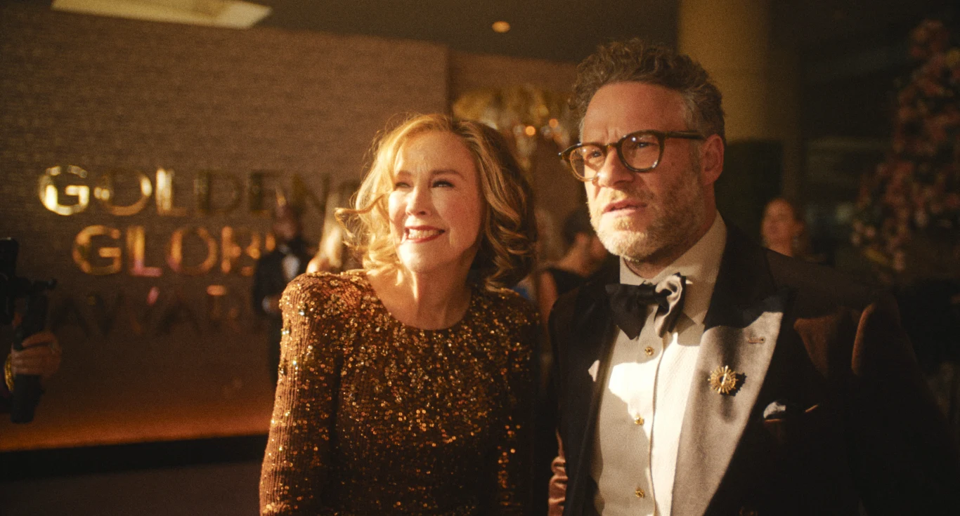 Catherine O'Hara i Seth Rogen w serialu "Studio" Elegancko ubrana kobieta w brokatowej sukni stoi obok mężczyzny w marynarce z muchą i okularach, oboje uśmiechają się, w tle złoty napis Golden Globe Awards oraz inni uczestnicy gali, oświetleni ciepłym światłem.