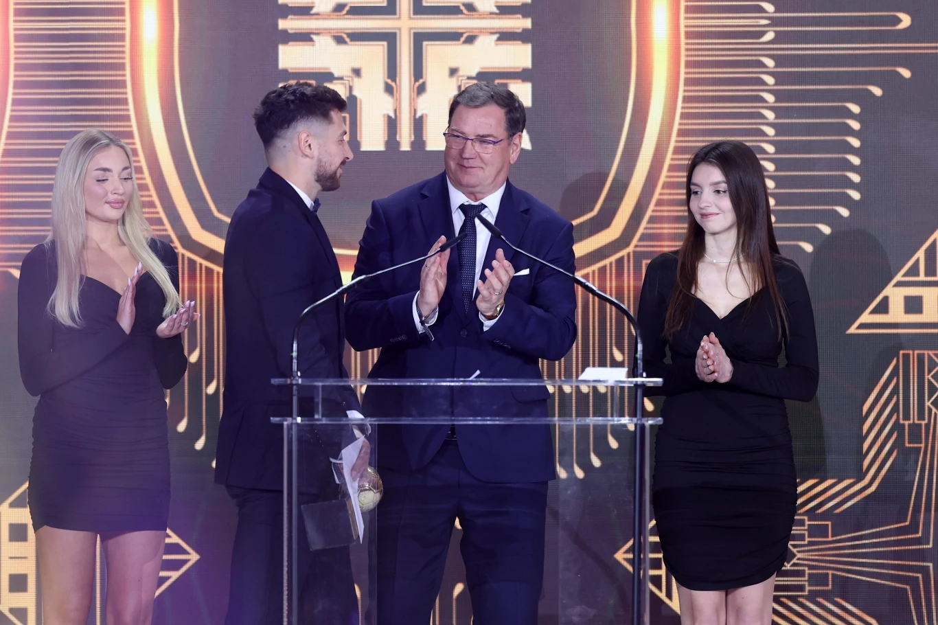 Cztery osoby w eleganckich strojach stoją na scenie za przezroczystą mównicą, dwie kobiety po bokach i dwóch mężczyzn pośrodku, jeden z nich trzyma statuetkę, wszyscy patrzą w stronę publiczności. Tło stanowi dekoracja sceniczna utrzymana w złotych i c...