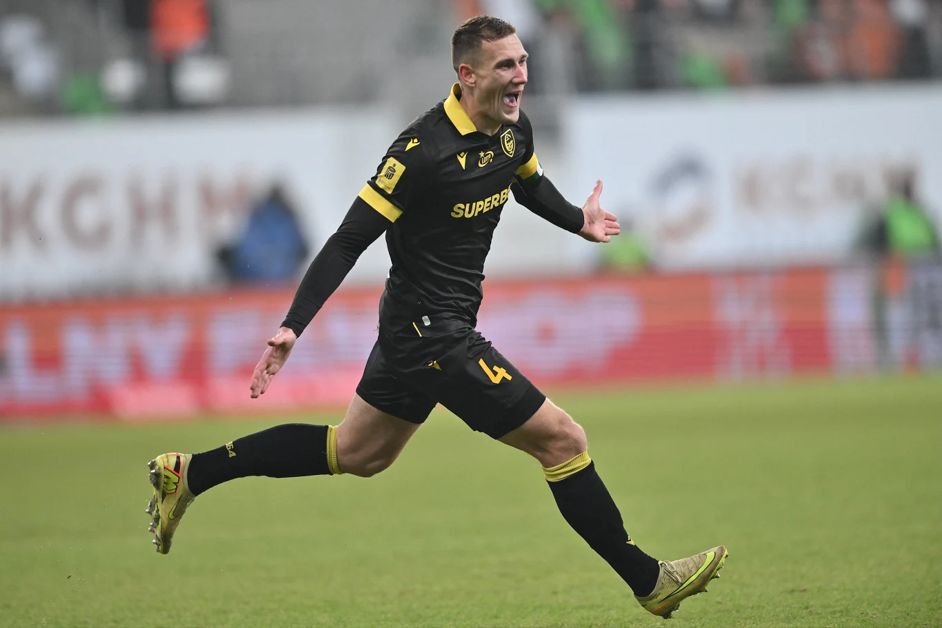 Piłkarz w czarnym stroju sportowym z numerem 4 dynamicznie biegnie po murawie z rozłożonymi ramionami, wyrażając radość, w tle widoczni są niescharakteryzowani kibice oraz reklamy stadionowe.