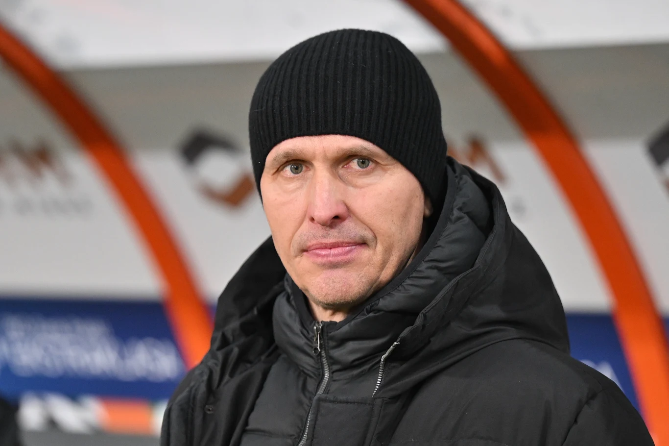 Mężczyzna w czarnej czapce oraz kurtce zimowej patrzy przed siebie, w tle widoczne rozmazane elementy stadionu.