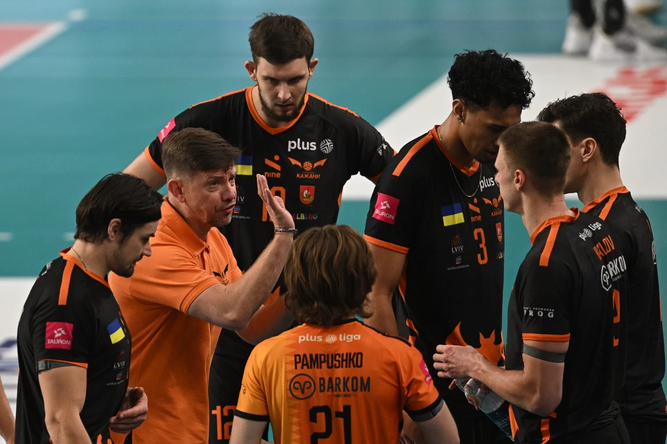 Trener drużyny siatkarskiej udziela wskazówek grupie zawodników ubranych w czarno-pomarańczowe stroje meczowe na tle boiska do siatkówki.