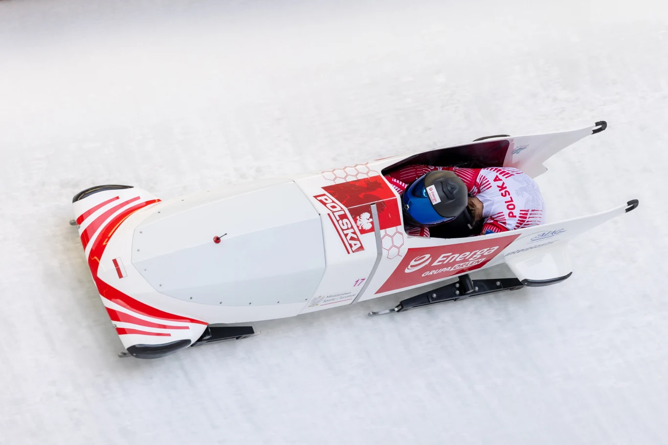 Biało-czerwony bobslej z polską flagą i oznaczeniami sponsorów pędzi po lodowej trasie, wewnątrz pojazdu dwóch zawodników w strojach sportowych i kaskach.