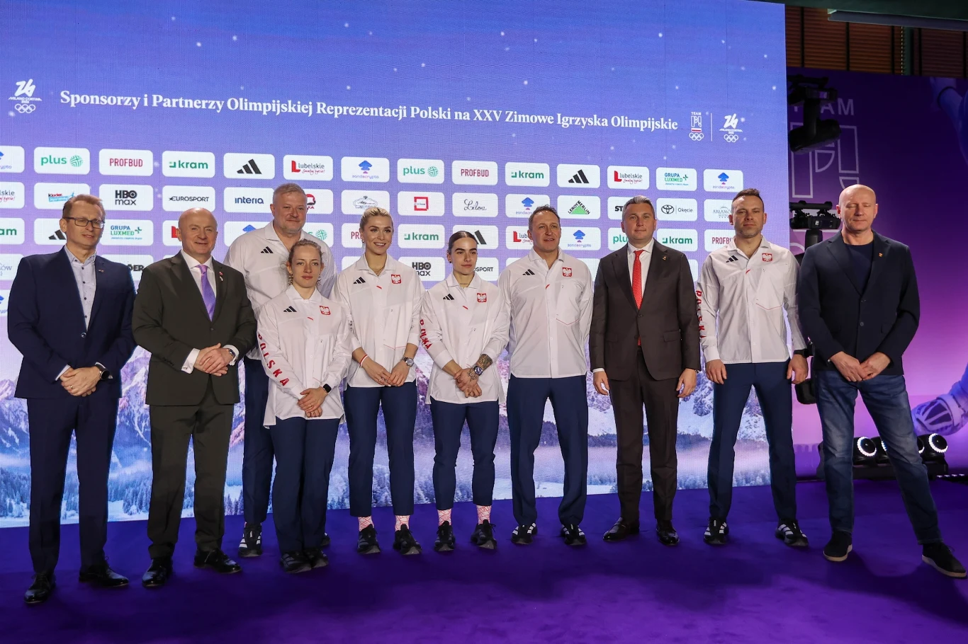 Grupa osób, w tym sportowców w białych sportowych kurtkach z logotypami oraz oficjalnie ubranych mężczyzn, stoi na tle ścianki z logotypami sponsorów i partnerów reprezentacji olimpijskiej. Uczestnicy patrzą w stronę obiektywu, w tle dominuje kolor fio...