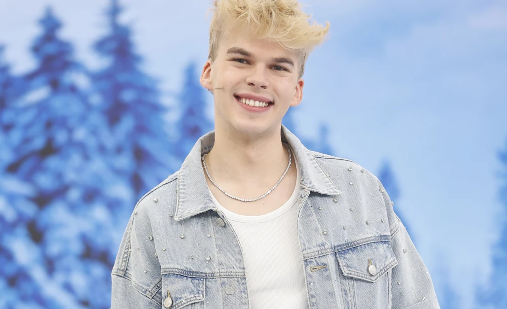 Młody mężczyzna z blond włosami szeroko się uśmiecha, ubrany jest w jasną, zdobioną kurtkę jeansową oraz biały t-shirt, tle rozmazowane sylwetki śnieżnych drzew.