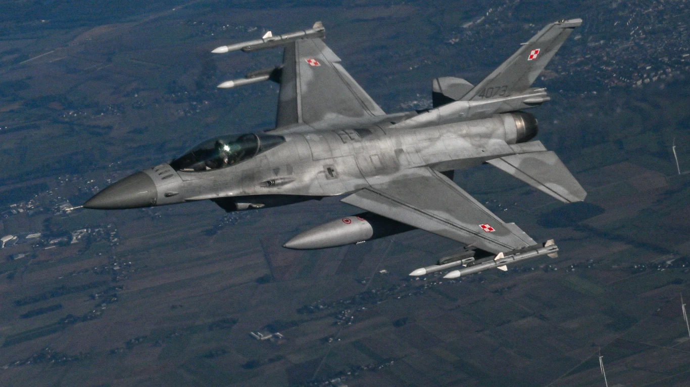 Nowoczesny myśliwiec F-16 z biało-czerwoną szachownicą lotnictwa wojskowego Polski leci nad rozległym, zielonym krajobrazem, uzbrojony w pociski powietrze-powietrze.