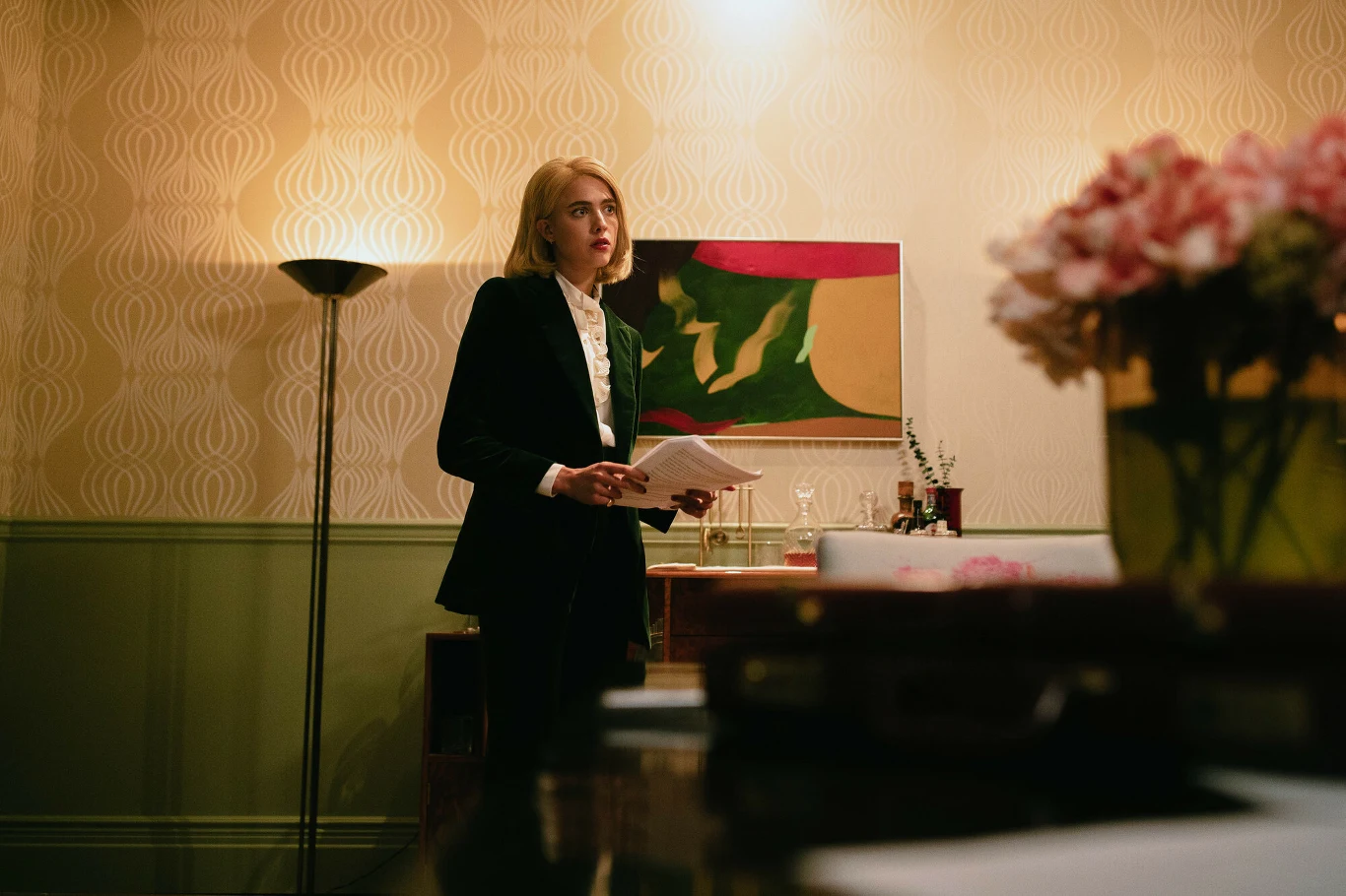 Młoda kobieta w garniturze stoi w eleganckim wnętrzu z dokumentami w rękach. W tle znajduje się nowoczesny obraz, lampa stojąca i komoda z karafką oraz szklankami. Na pierwszym planie widoczny jest bukiet kwiatów.