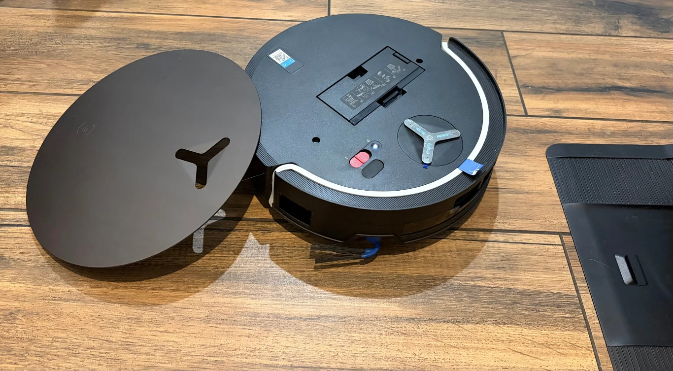 Okrągły robot sprzątający z otwartą pokrywą górną ukazuje wnętrze urządzenia, leży na drewnianej podłodze obok czarnego elementu wyposażenia.