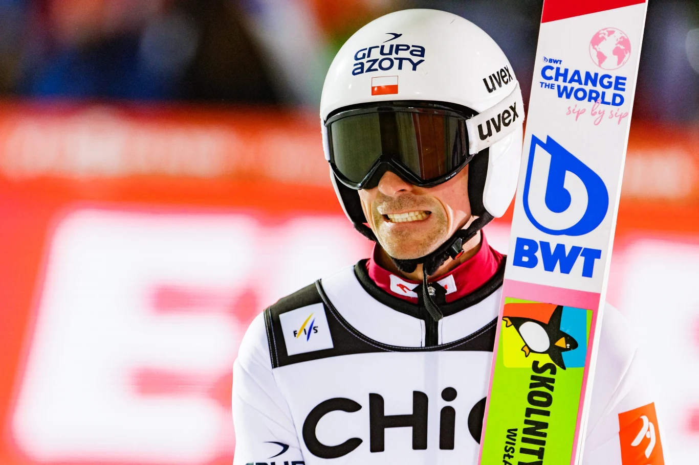 Piotr Żyła Skoczek narciarski w kombinezonie startowym i kasku, trzymający narty z widocznymi logotypami sponsorów, w tle rozmyte światła stadionu.