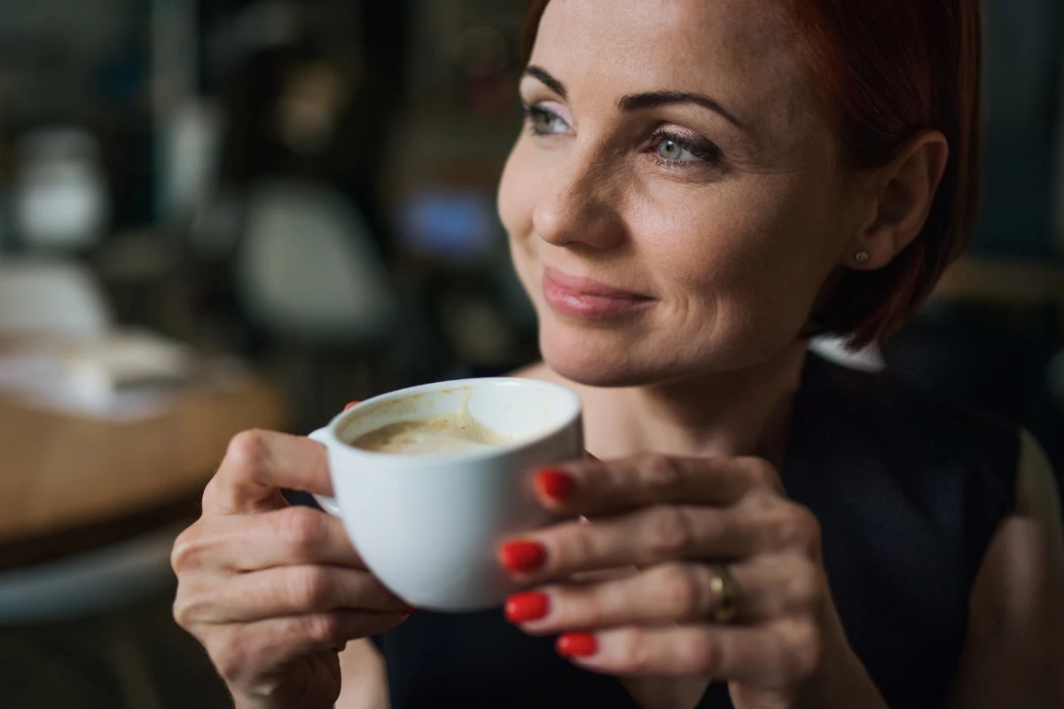 Jedna łyżeczka w kawie może wspierać naturalne procesy anti-aging organizmu Kobieta z krótkimi, rudymi włosami trzyma filiżankę kawy oburącz i uśmiecha się lekko patrząc na bok. Jej paznokcie są pomalowane na czerwono. W tle rozmazane wnętrze kawiarni.