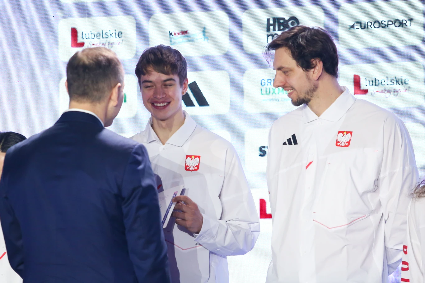 Władimir Semirunnij odbierający nominację olimpijską od prezydenta RP Karola Nawrockiego Trzech mężczyzn ubranych w białe stroje reprezentacji Polski znajduje się na tle tablicy z logotypami sponsorów, dwaj z nich mają naszywkę z orłem na piersi, a jeden uśmiecha się, trzymając dokument.
