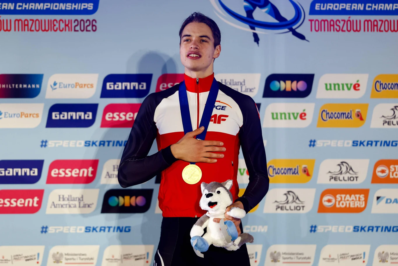 Władimir Semirunnij wystartuje na ZIO w Mediolanie na dwóch dystansach: 1500 m i 10000 m. Wciąż ma szansę także na 5000 m Sportowiec w stroju reprezentacyjnym stoi na podium z zawieszoną na szyi złotą medalą, trzymając pluszową maskotkę, na tle baneru z logotypami sponsorów i nazwą zawodów.