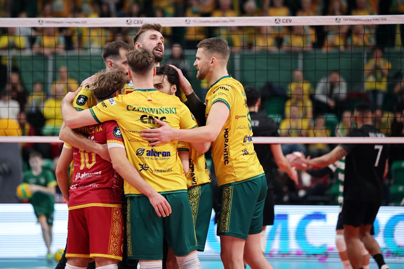 Grupa siatkarzy w żółto-zielonych strojach świętuje zdobycie punktu na boisku, obejmując się grupowo, w tle tłum kibiców na trybunach.