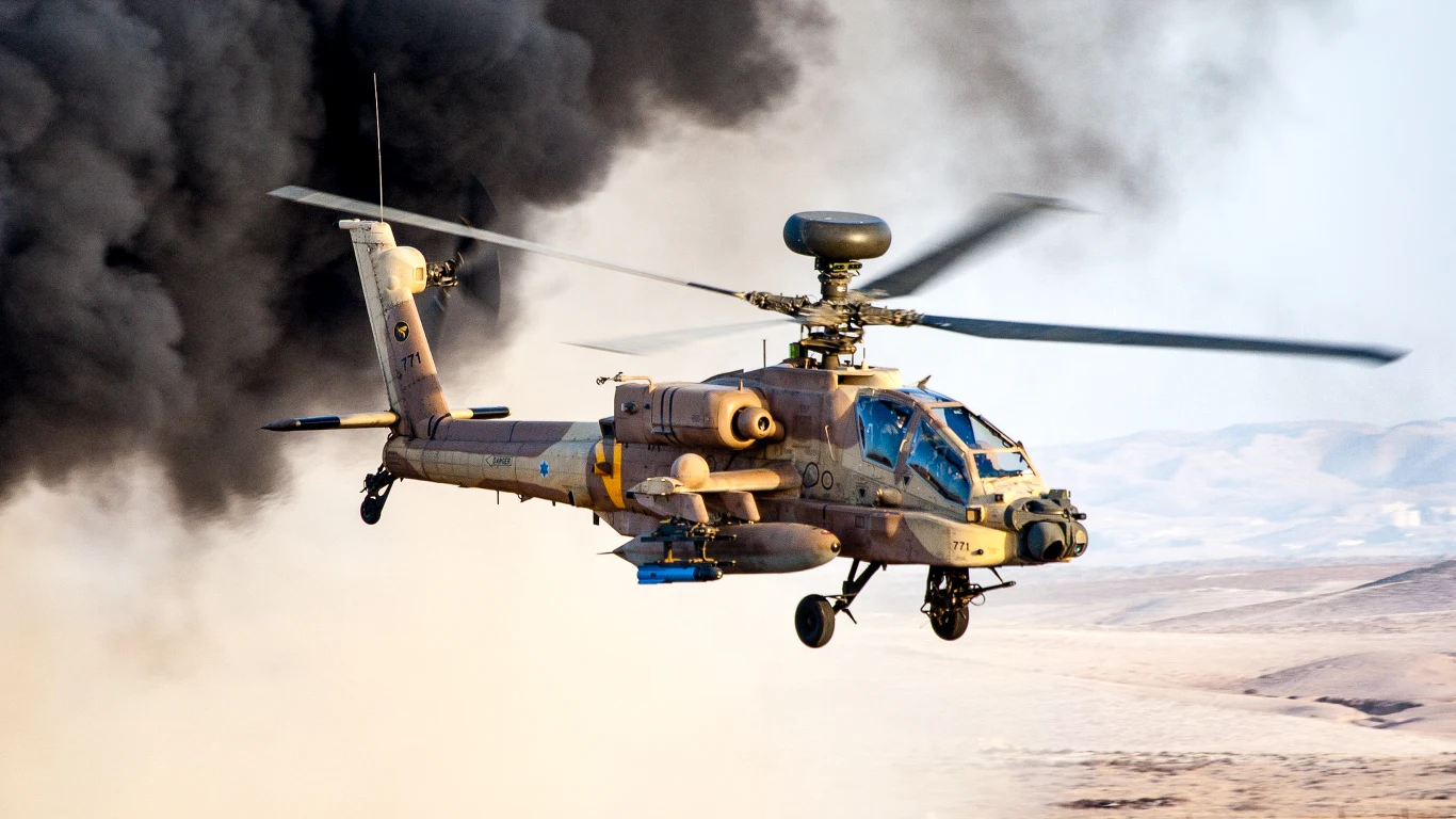 Izrael chce kupić od Amerykanów najnowsze Apache Śmigłowiec bojowy Apache unosi się nad pustynnym terenem, w tle gęste chmury czarnego dymu i pagórkowaty krajobraz.