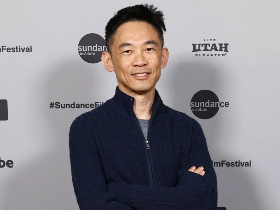 James Wan Mężczyzna w granatowym swetrze stoi z założonymi rękami na tle ścianki z logotypami festiwalu filmowego.