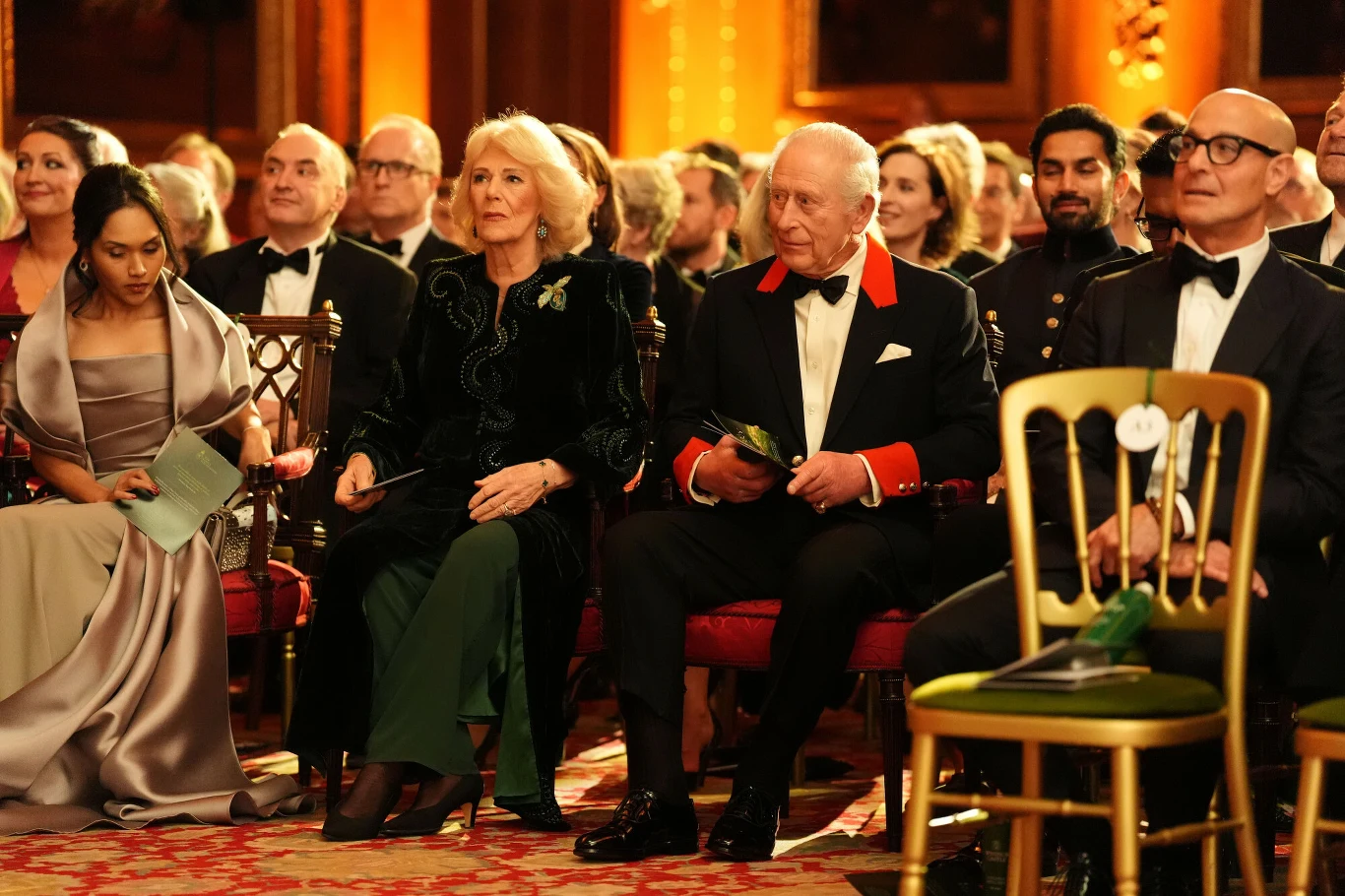 Grupa elegancko ubranych osób siedzi w rzędzie podczas uroczystego wydarzenia w bogato zdobionej sali z czerwonym dywanem. Na pierwszym planie mężczyzna w smokingu i kobieta w zielonej marynarce, wokół nich pozostali uczestnicy.